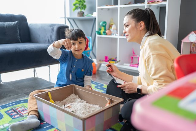 Child plays with box of sand next to a woman with a clipboard