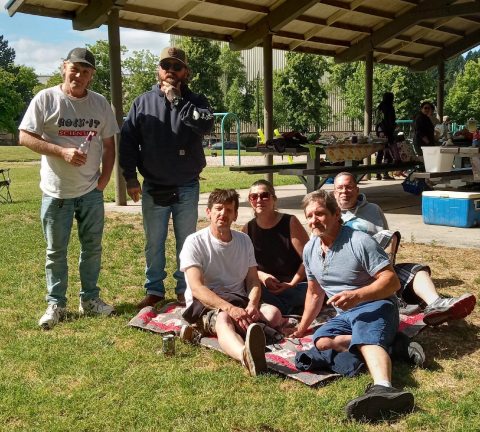 A group of men pose on a picnic blanket with a BBQ in the background