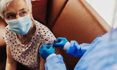 White haired woman in face mask has bandage applied to her arm by a healthcare provider in blue gloves and surgical gown after getting a shot or vaccine 