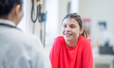 Close-up of the back of a doctor during a visit with a smiling young teen wearing a red shirt