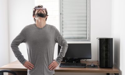 Bearded man in long-sleeve gray shirt looks up to ceiling, stretching back in front of desk