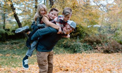 Three young children ride piggyback on a smiling red-haired man outside with fall leaves on the ground 