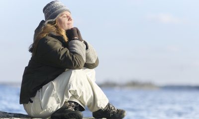 Photograph of adult white woman in knit hat and parka sitting cross-legged on a beach and meditating.