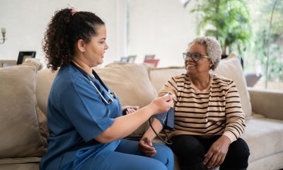 Dark-haired healthcare provider in blue scrubs takes blood pressure reading of older Black patient sitting on couch