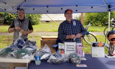 Two men stand behind a table filled with bicycle helmets.