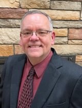 Photo of Faye wearing a black suit with a red shirt and tie in front of a brick building