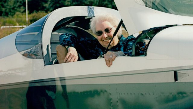 An elderly woman wearing sunglasses smiles from inside a small plane.