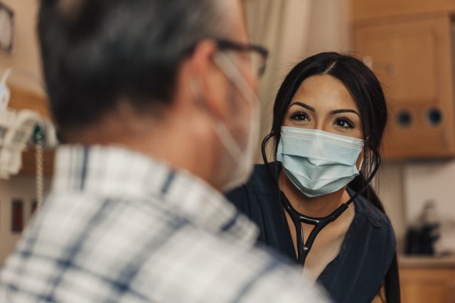 A female nurse wearing a surgical mask works with a male patient also wearing a mask.