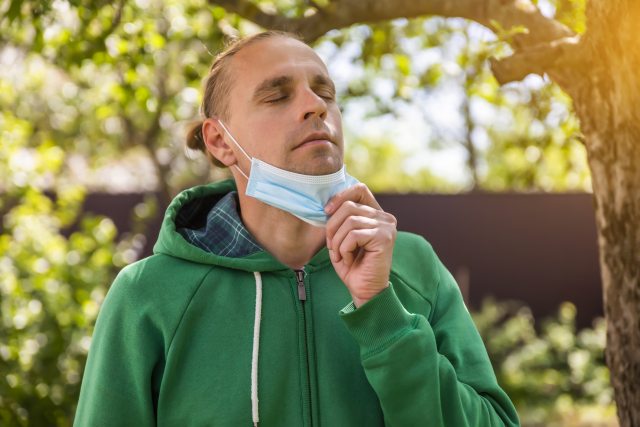 White man in a park wearing a green hoodie removes his medical mask to breathe clean air.