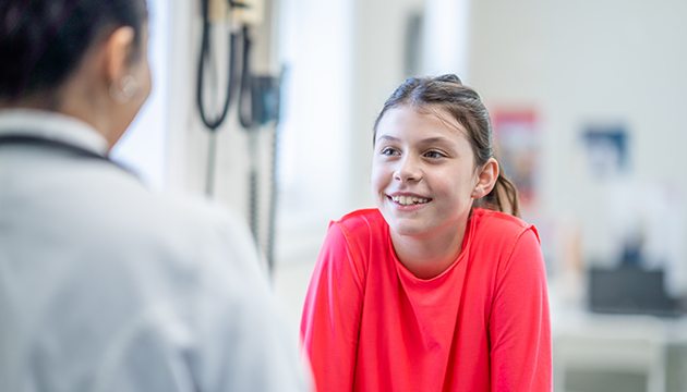 Close-up of the back of a doctor during a visit with a smiling young teen wearing a red shirt
