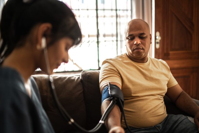 Young healthcare provider takes blood pressure reading for a Black man sitting on a couch