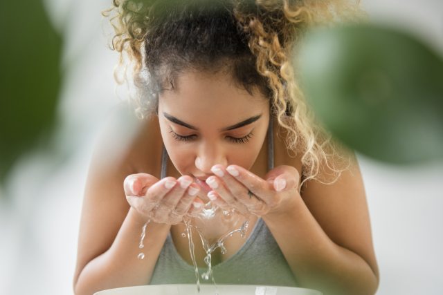 Close up of a woman rinsing her face with water. She has light brown curly hair pulled back from her face and is wearing a green tank top.