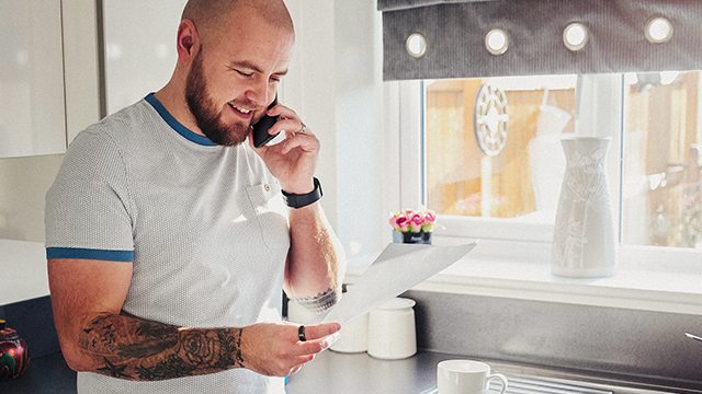 An adult male, standing in a well-lit kitchen, smiles and looks over a bill while speaking on the phone