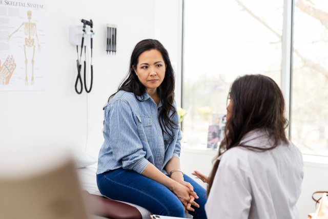 Black-haired woman in a blue shirt and jeans sits on an exam tableand listens as her female doctor shares information.