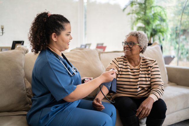 Dark-haired healthcare provider in blue scrubs takes blood pressure reading of older Black patient sitting on couch